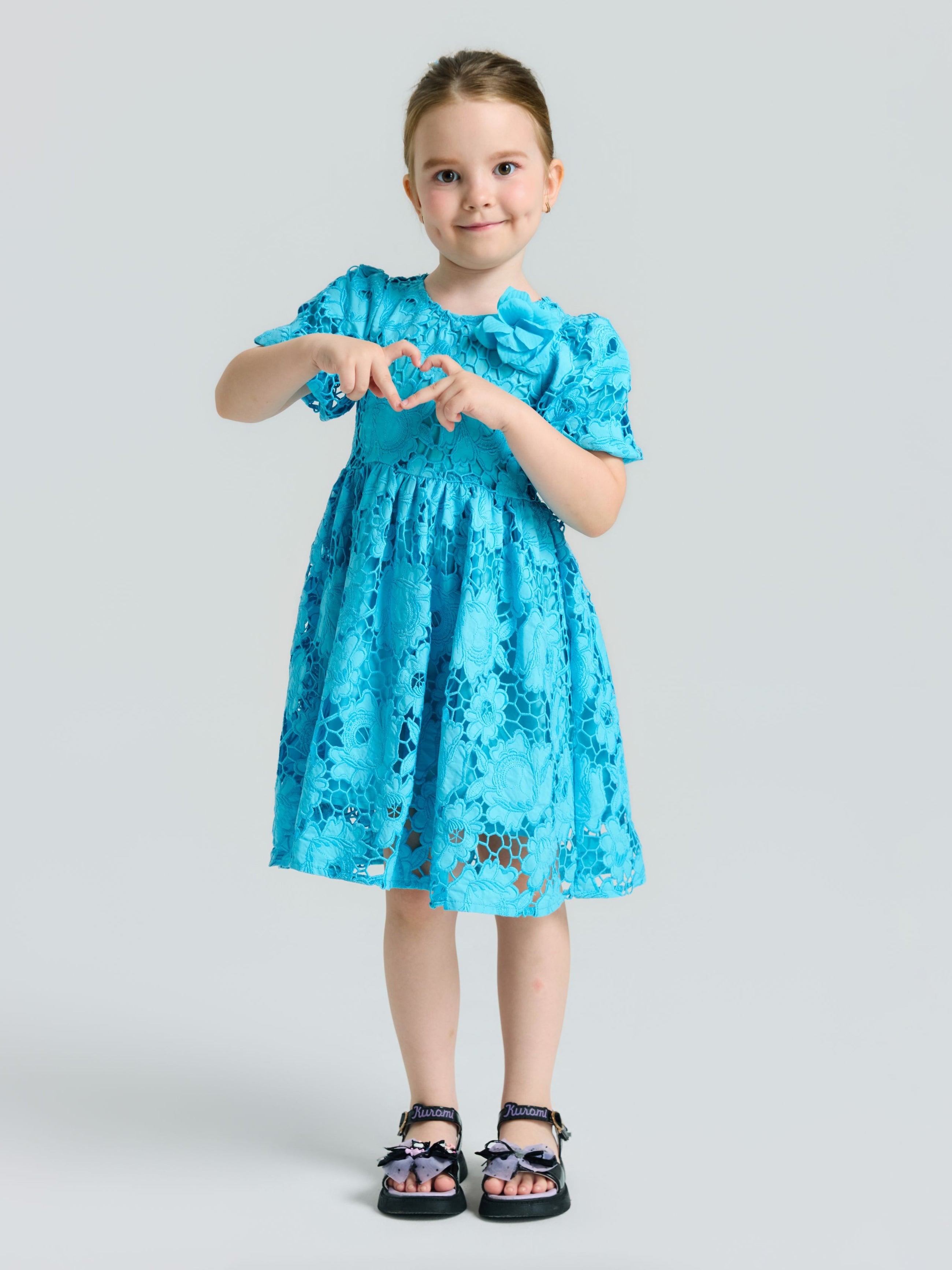 A young girl wearing the Social Queen Girls 3D Floral Appliqué Cutwork Lace Dress and sandals stands against a plain background, forming a heart shape with her hands.