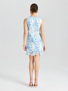 A woman faces away in the Social Queen Embellished Floral Lace Mini Dress, featuring sleeveless light blue lace with organza flowers and paired with beige heels, set against a plain white background.