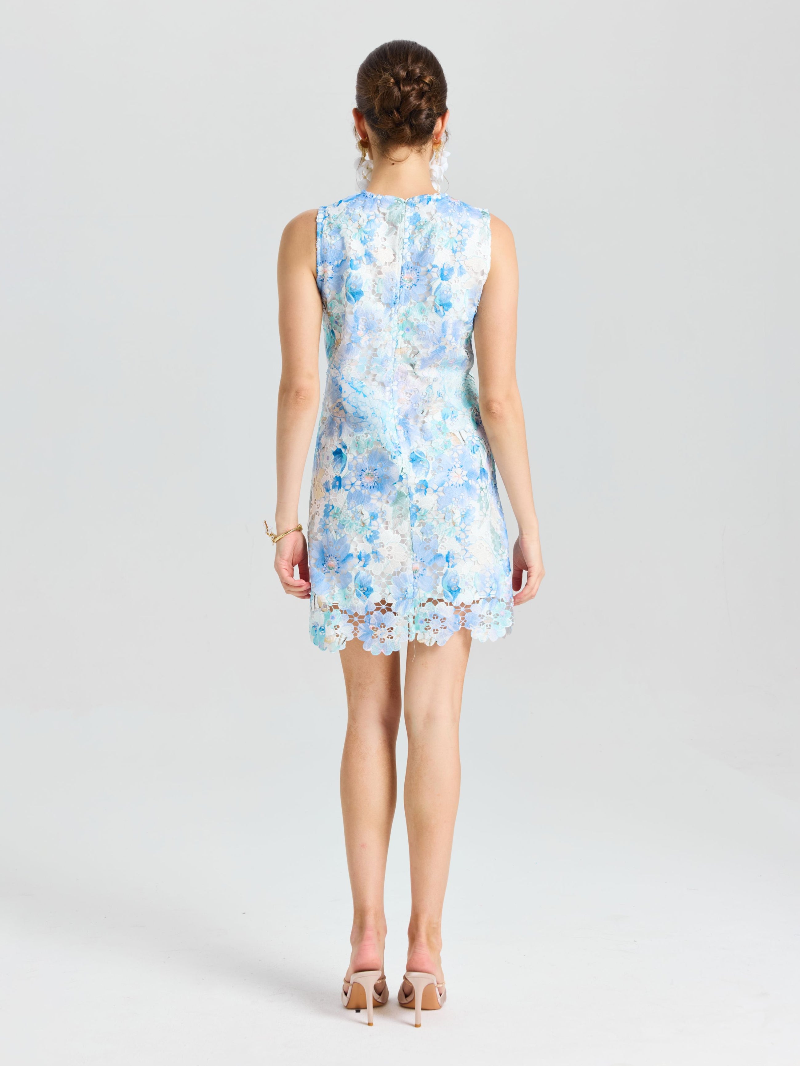 A woman faces away in the Social Queen Embellished Floral Lace Mini Dress, featuring sleeveless light blue lace with organza flowers and paired with beige heels, set against a plain white background.