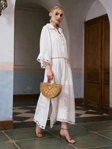 A woman stands indoors wearing the Social Queen Floral Cutwork Embroidered Linen Dress with gold heels, holding a semi-circular bamboo handbag.