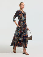 A woman stands against a plain background wearing the Social Queen Paisley Print Knit Dress (Belt Included), styled with three-quarter sleeves, sandals, and a small handbag.