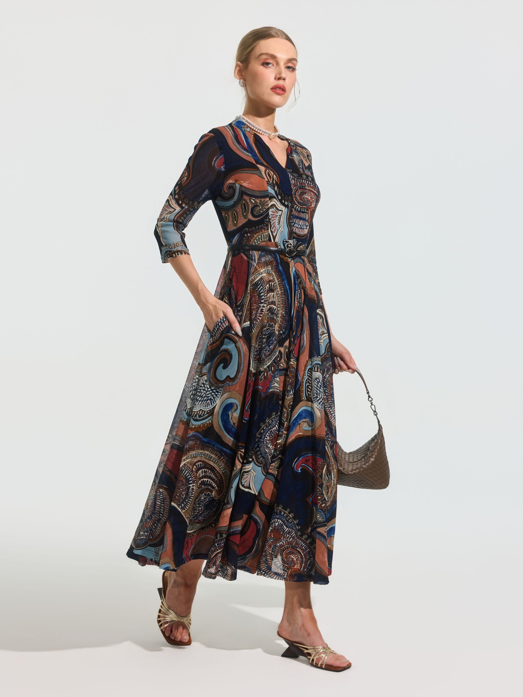 A woman stands against a plain background wearing the Social Queen Paisley Print Knit Dress (Belt Included), styled with three-quarter sleeves, sandals, and a small handbag.