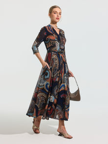 A woman stands against a plain background wearing the Social Queen Paisley Print Knit Dress (Belt Included), styled with three-quarter sleeves, sandals, and a small handbag.