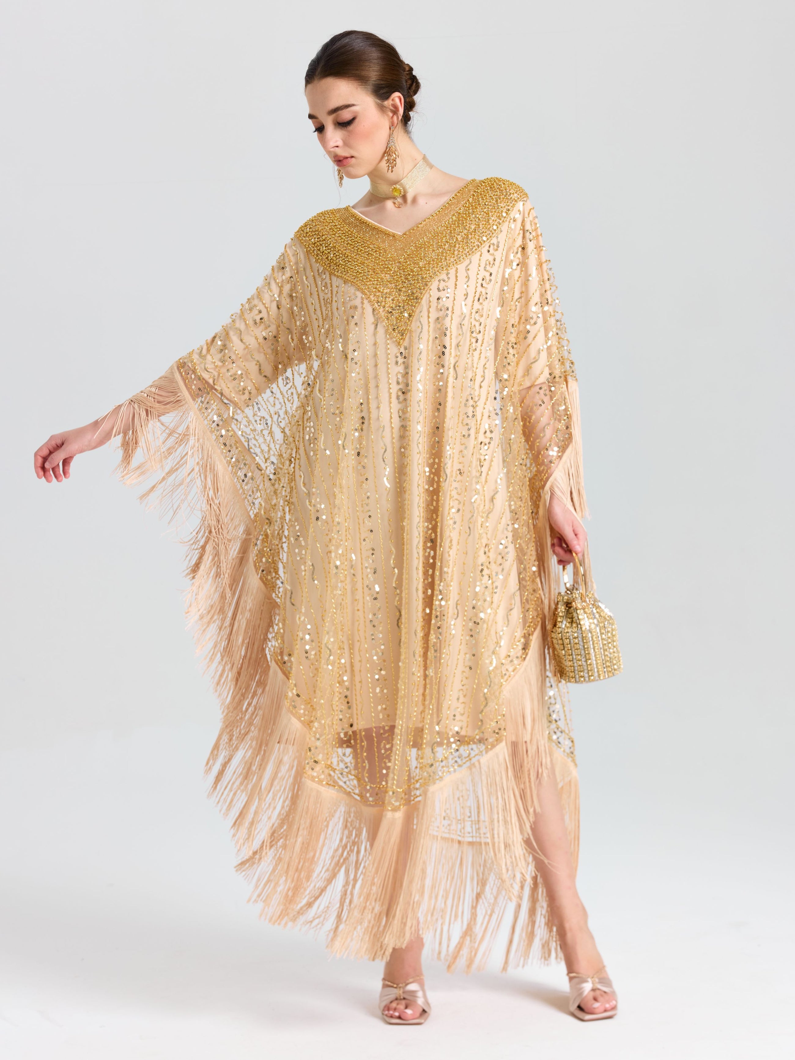 A woman wears the Social Queen Beaded Fringe Cape Dress with sequin embellishments, paired with a beaded purse, matching earrings, and heels, standing against a plain light background.
