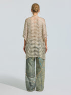 A person stands facing away, styled in a neat bun and wearing the Social Queen Pearl Daisy Cutwork Long Cardigan—with sheer grid patterns and subtle pearl accents—layered over loose, light blue jeans with decorative designs.
