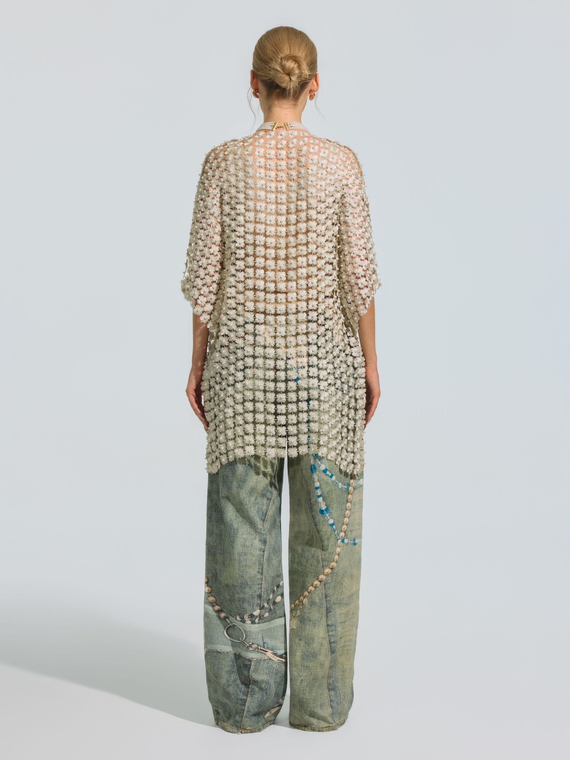 A person stands facing away, styled in a neat bun and wearing the Social Queen Pearl Daisy Cutwork Long Cardigan—with sheer grid patterns and subtle pearl accents—layered over loose, light blue jeans with decorative designs.