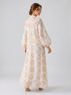 A woman stands in profile against a plain light background, wearing the Social Queen Leaf Embroidered Beaded Dress—a long, white dress featuring tan leaf embroidery and balloon sleeves—paired with beige high-heeled shoes.