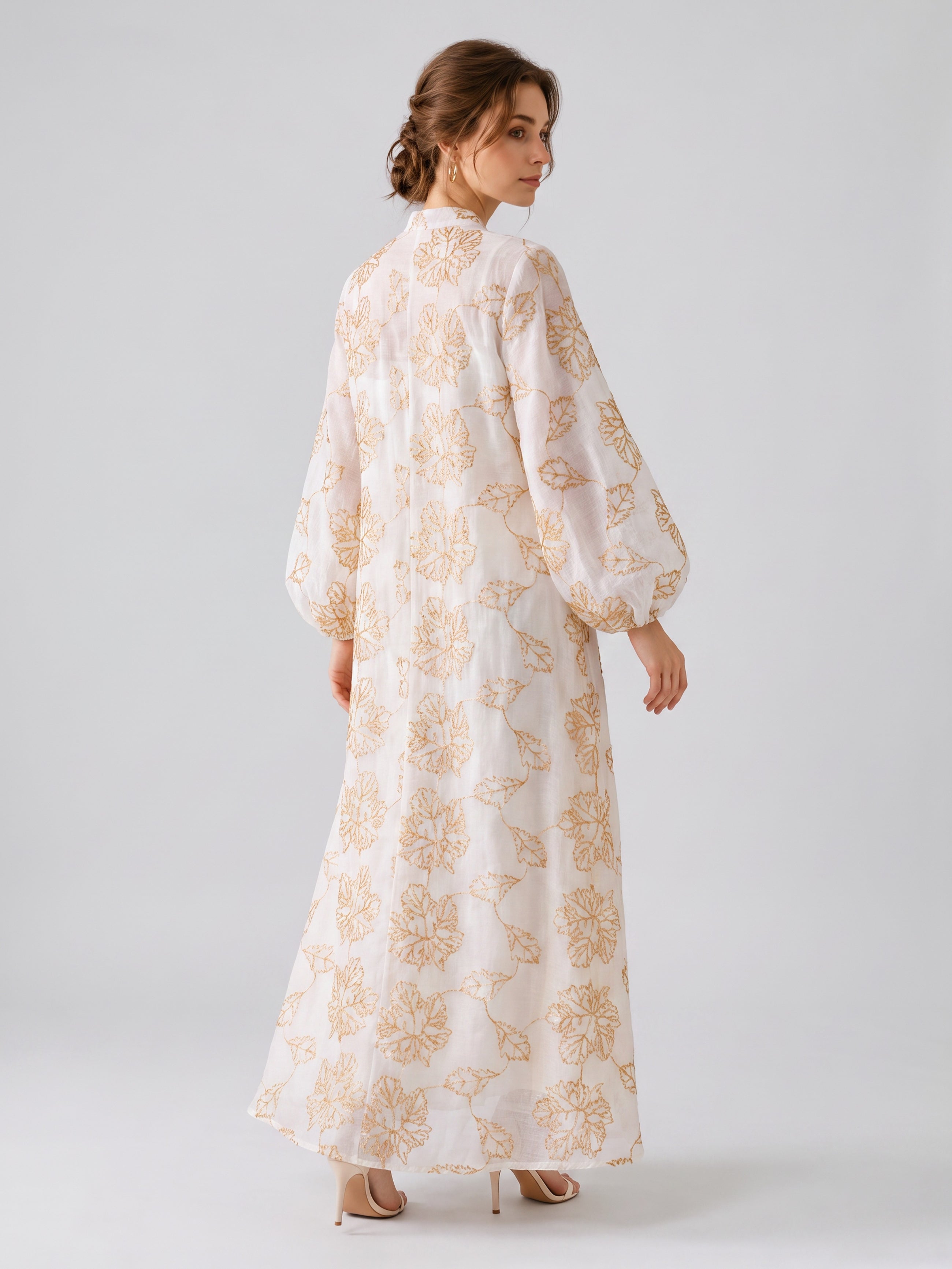 A woman stands in profile against a plain light background, wearing the Social Queen Leaf Embroidered Beaded Dress—a long, white dress featuring tan leaf embroidery and balloon sleeves—paired with beige high-heeled shoes.