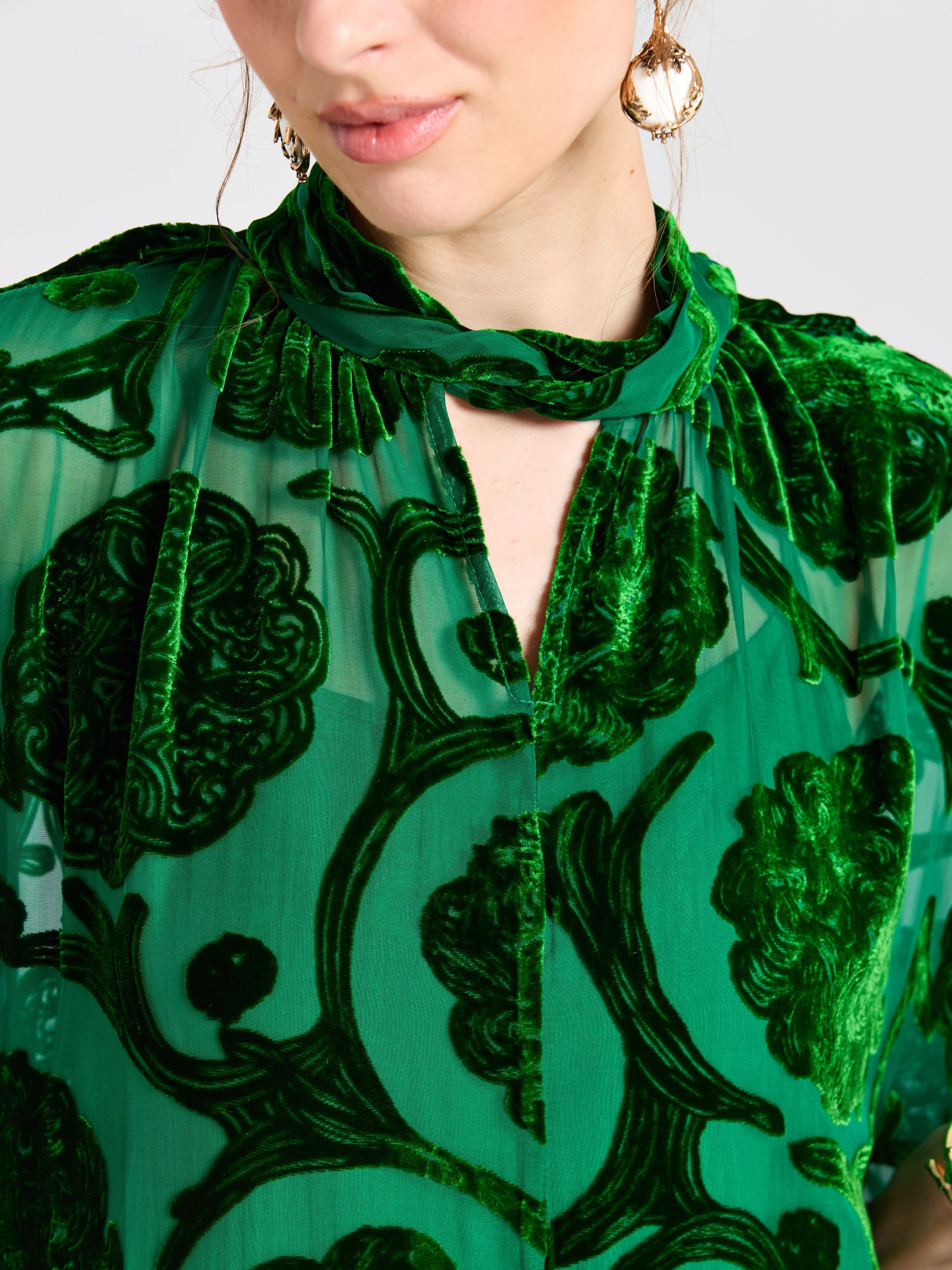 A woman in the Floral Velvet Cape Dress (Cami Included) by Social Queen, a sheer green style with a floral burnout pattern, paired with a matching scarf and gold hoop earrings.