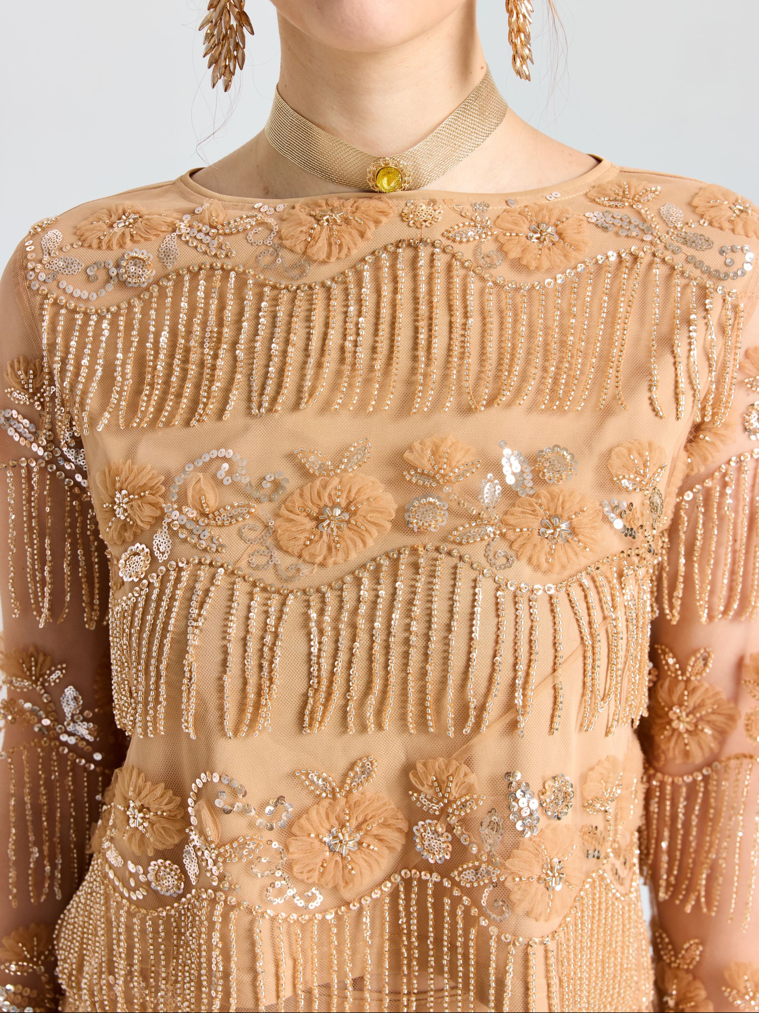 A person wearing the Beaded Fringe Floral Top by Social Queen, featuring beige sheer mesh with beadwork, sequins, floral embroidery, and beaded fringe, styled with a gold choker and dangling earrings.