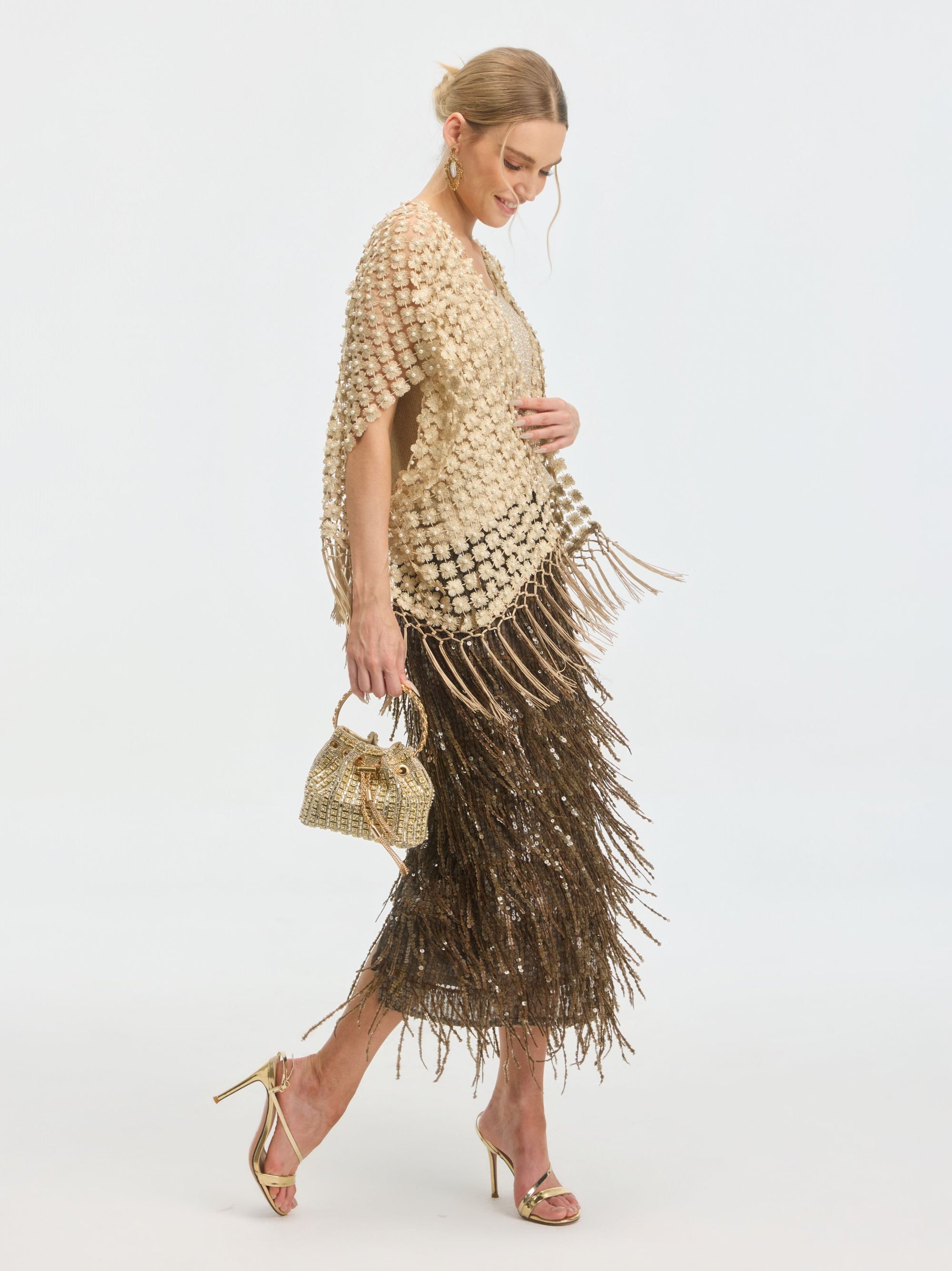 A woman in gold heels and a textured cream top wears Social Queen's Pearl Floral Appliqué Fringe Cape over a bronze fringe skirt, holding a small gold handbag, posing against a plain white background.
