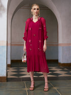 A woman stands indoors in front of an arched doorway, holding a small handbag and wearing strappy heels, styled in the Social Queen 3D Floral Pearl Mulberry Silk Dress—a knee-length, long-sleeved magenta dress adorned with intricate 3D floral appliqué.