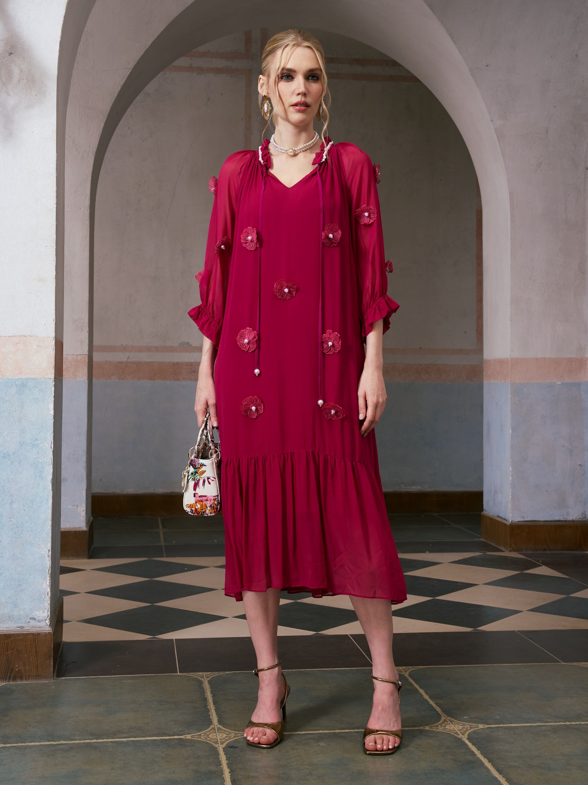A woman stands indoors in front of an arched doorway, holding a small handbag and wearing strappy heels, styled in the Social Queen 3D Floral Pearl Mulberry Silk Dress—a knee-length, long-sleeved magenta dress adorned with intricate 3D floral appliqué.