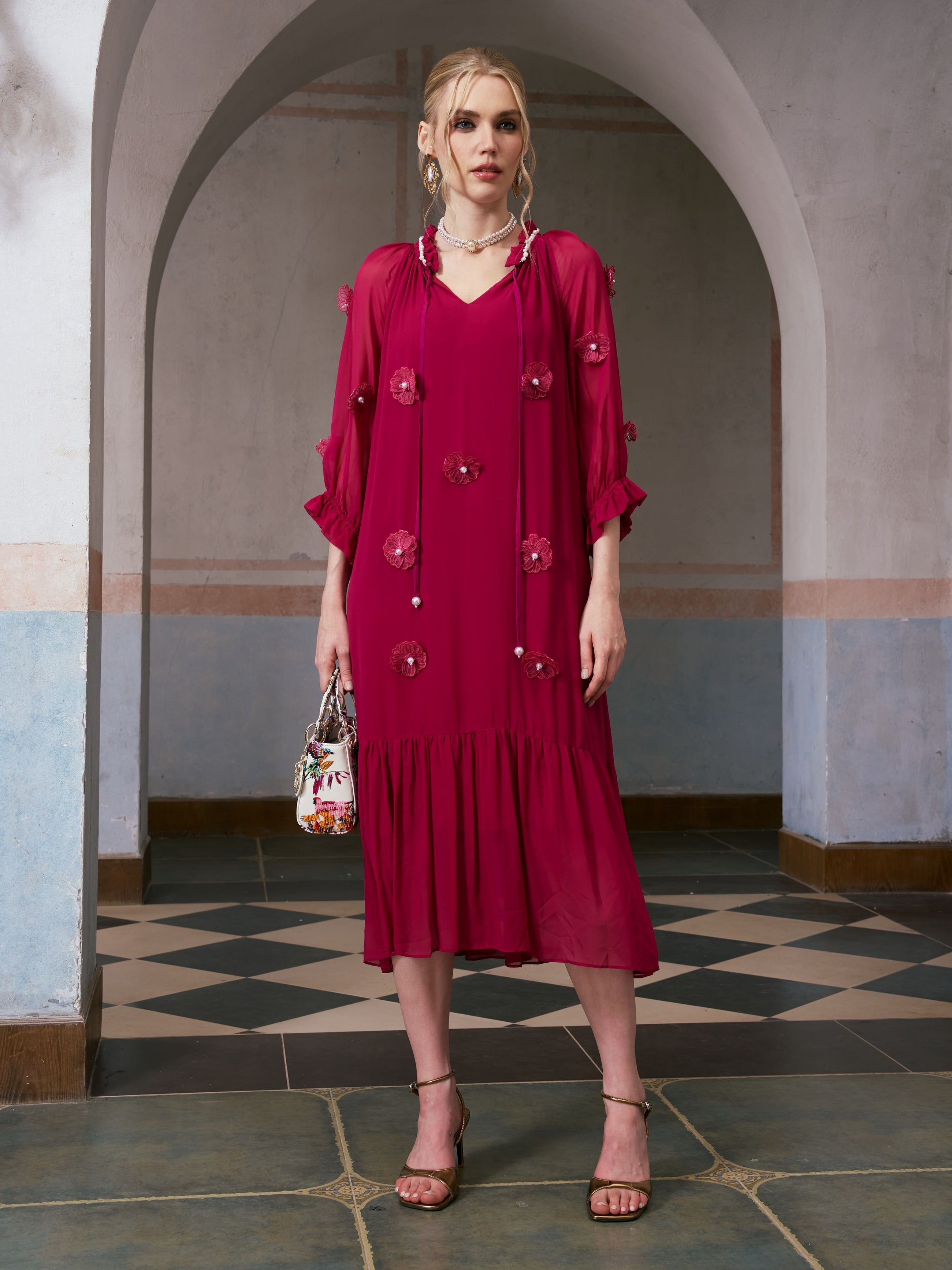 A woman stands indoors in front of an arched doorway, holding a small handbag and wearing strappy heels, styled in the Social Queen 3D Floral Pearl Mulberry Silk Dress—a knee-length, long-sleeved magenta dress adorned with intricate 3D floral appliqué.