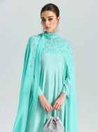 A woman in Social Queen’s Beaded Cape Scarf Gown with a matching purse stands against a plain background, showcasing the ideal elegant wedding guest look.