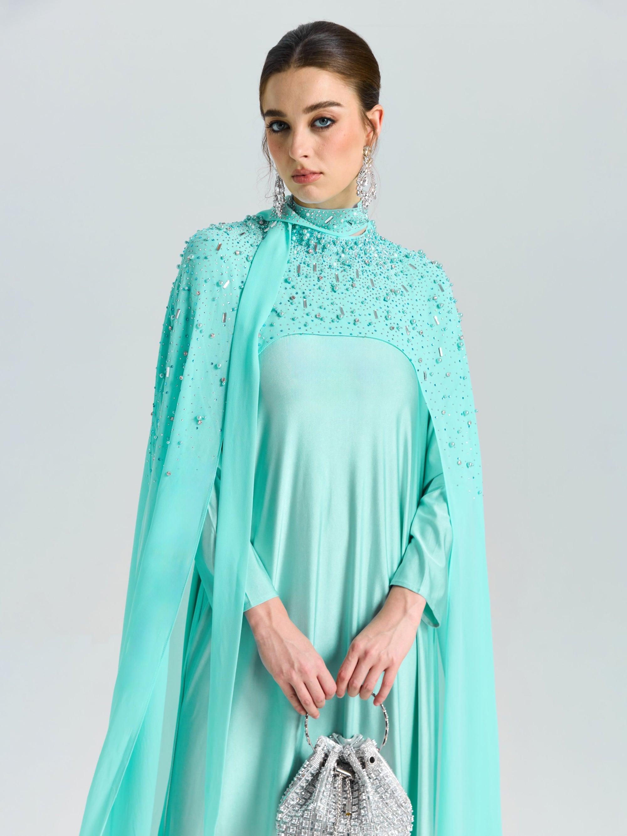 A woman in Social Queen’s Beaded Cape Scarf Gown with a matching purse stands against a plain background, showcasing the ideal elegant wedding guest look.