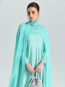 A woman in Social Queen’s Beaded Cape Scarf Gown with a matching purse stands against a plain background, showcasing the ideal elegant wedding guest look.
