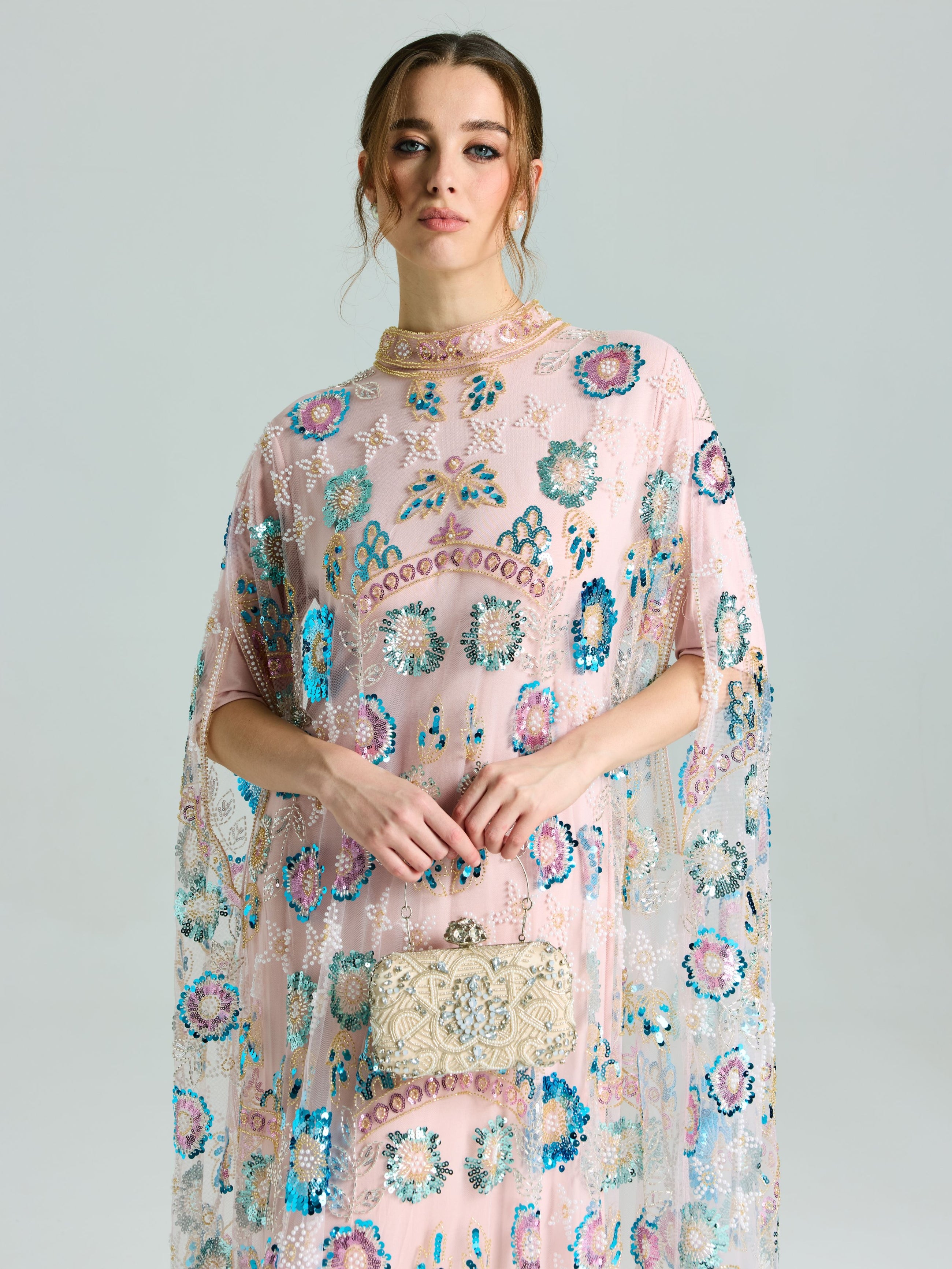 A woman stands against a plain background, wearing the Social Queen Beaded Floral Embellished Cape Gown, and holding a small ornate clutch bag.