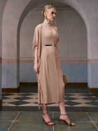 A woman stands on a checkered floor in front of an arched doorway, holding a wicker bag and wearing the Social Queen Asymmetric Fringe Midi Dress (belt included) in beige, paired with gold heels.