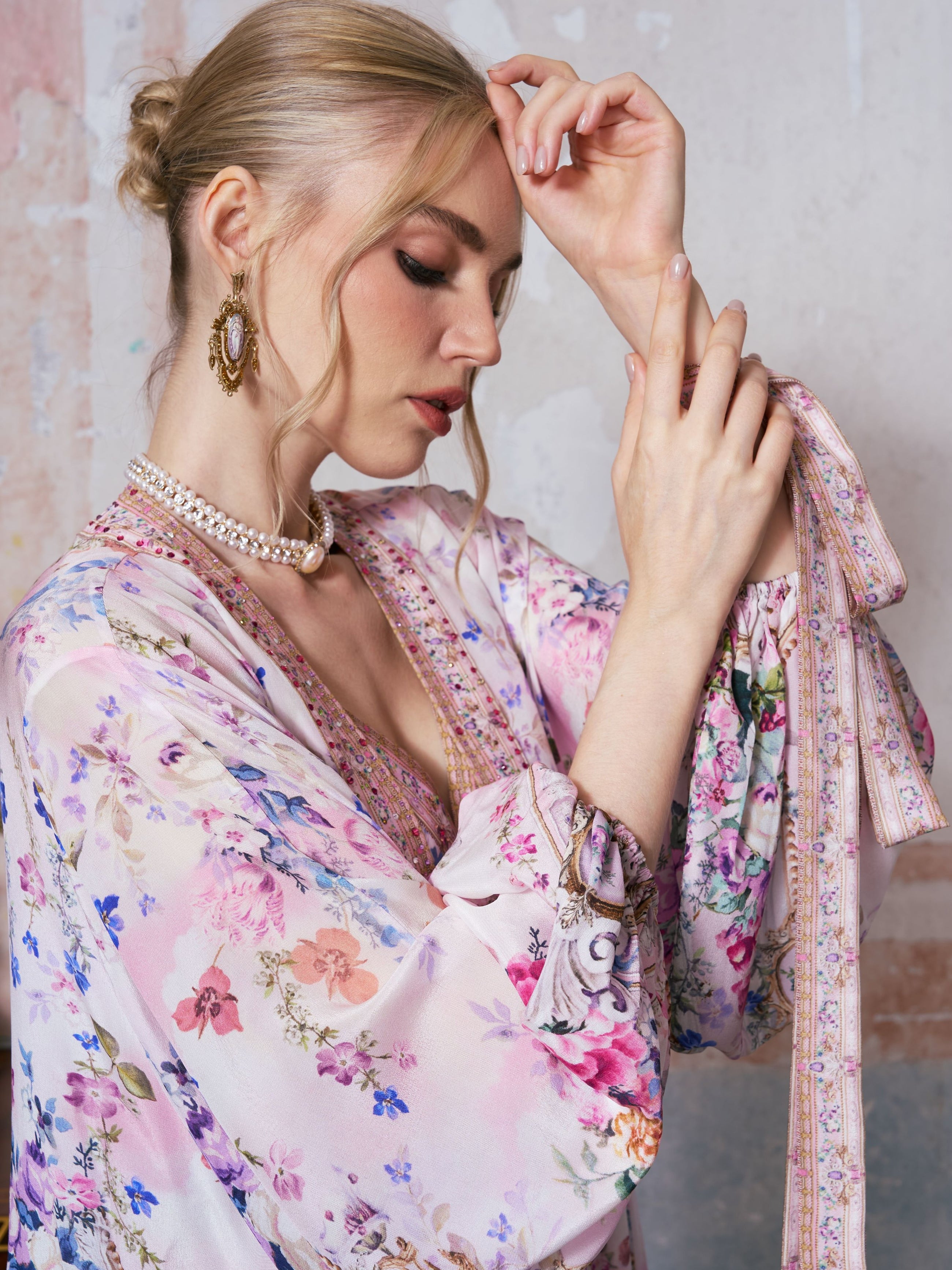 A woman with blonde hair in a bun wears the Social Queen Baroque Print Mulberry Silk Robe with Scarf, paired with a pearl necklace and large earrings, gazing down as she raises one arm and touches her wrist against a neutral background.