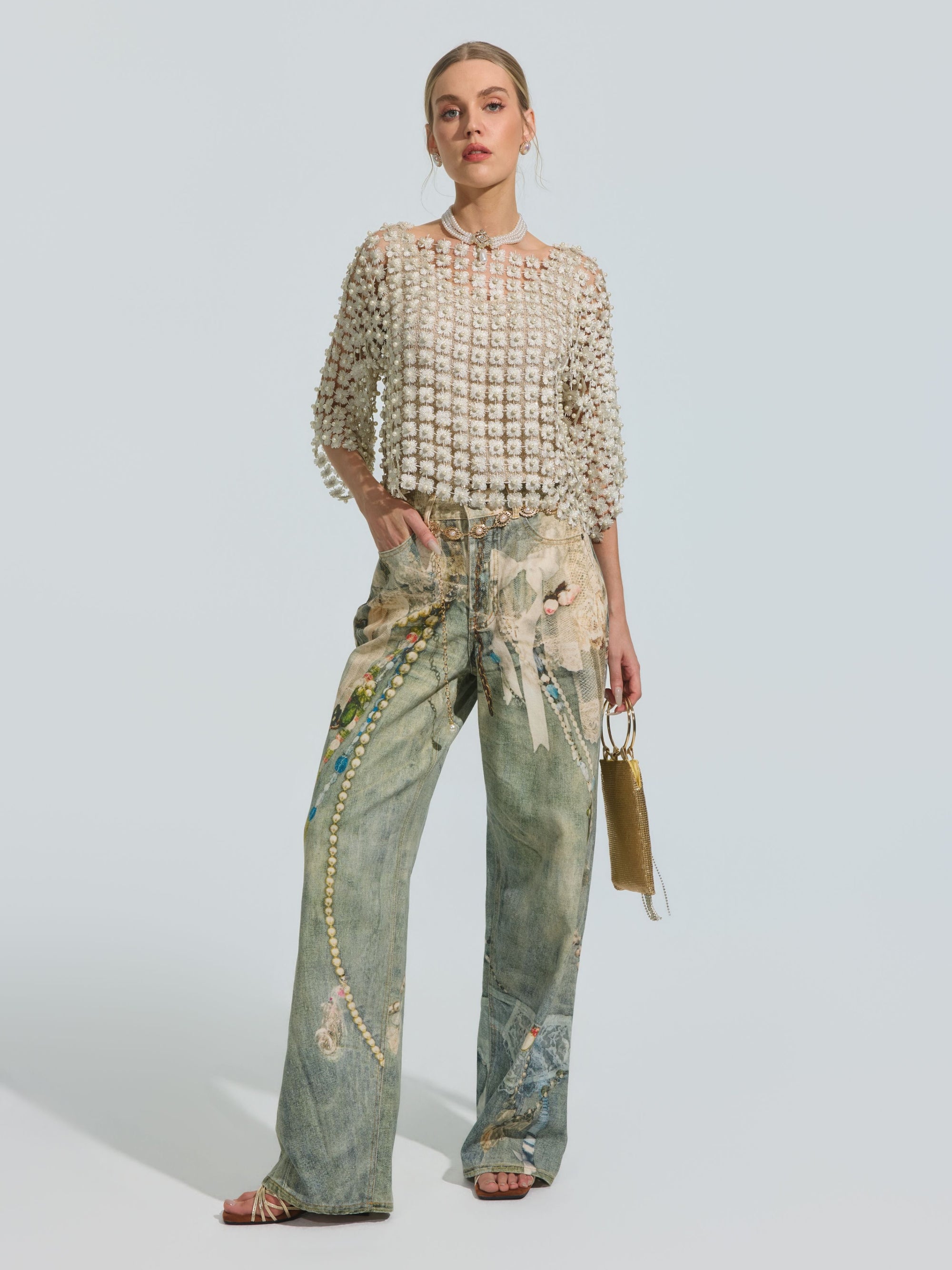 A woman stands facing forward against a plain light background, wearing Social Queen’s Pearl Chain Printed Jeans in a wide-leg silhouette with embellished details, paired with a beaded sheer top, sandals, and holding a small gold handbag.