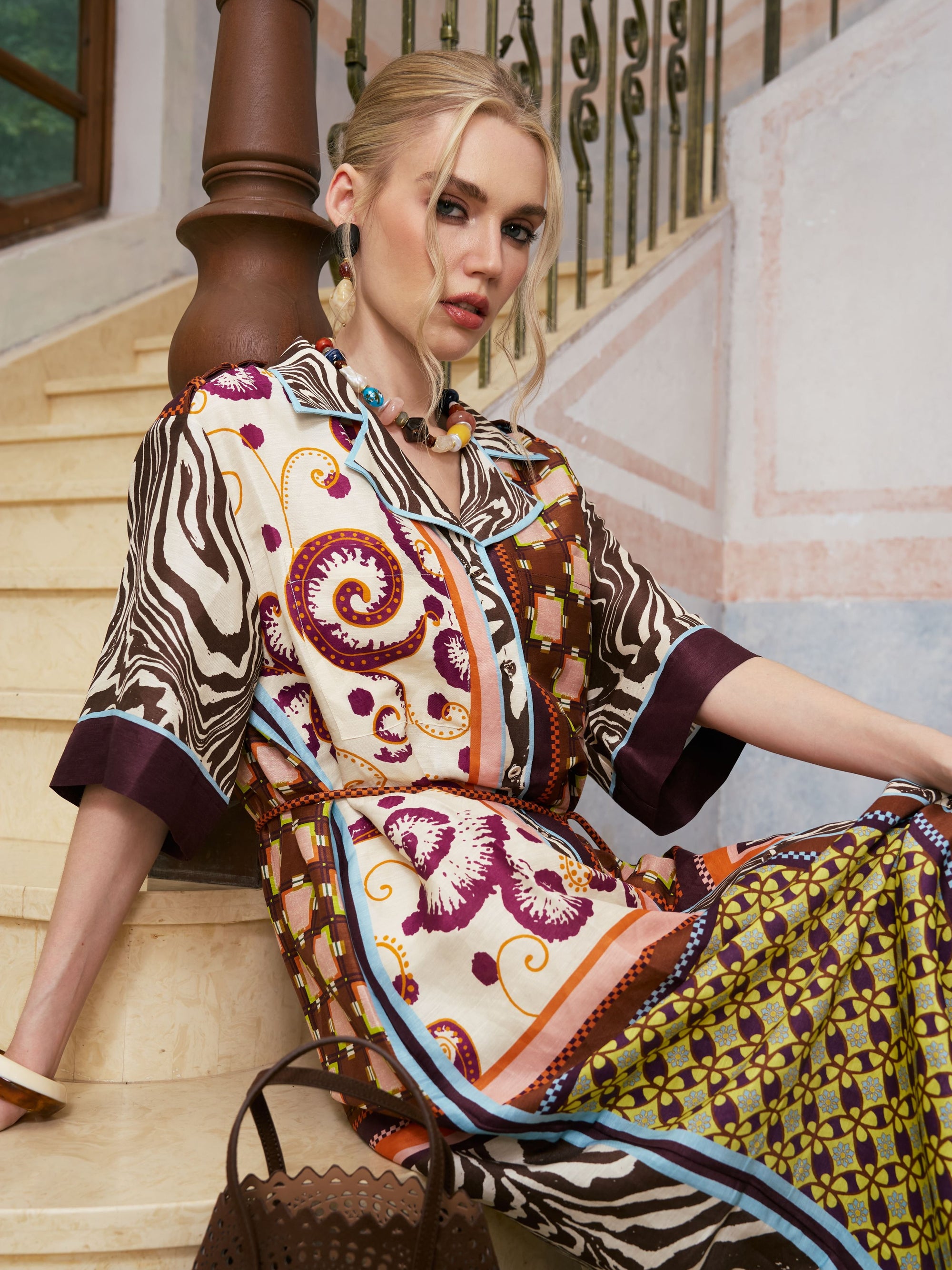A woman in the Social Queen Patchwork Print Cotton Shirt Dress (belt included) sits on marble stairs, leaning against a wooden banister, with a brown handbag on the step beside her.