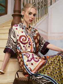 A woman in the Social Queen Patchwork Print Cotton Shirt Dress (belt included) sits on marble stairs, leaning against a wooden banister, with a brown handbag on the step beside her.