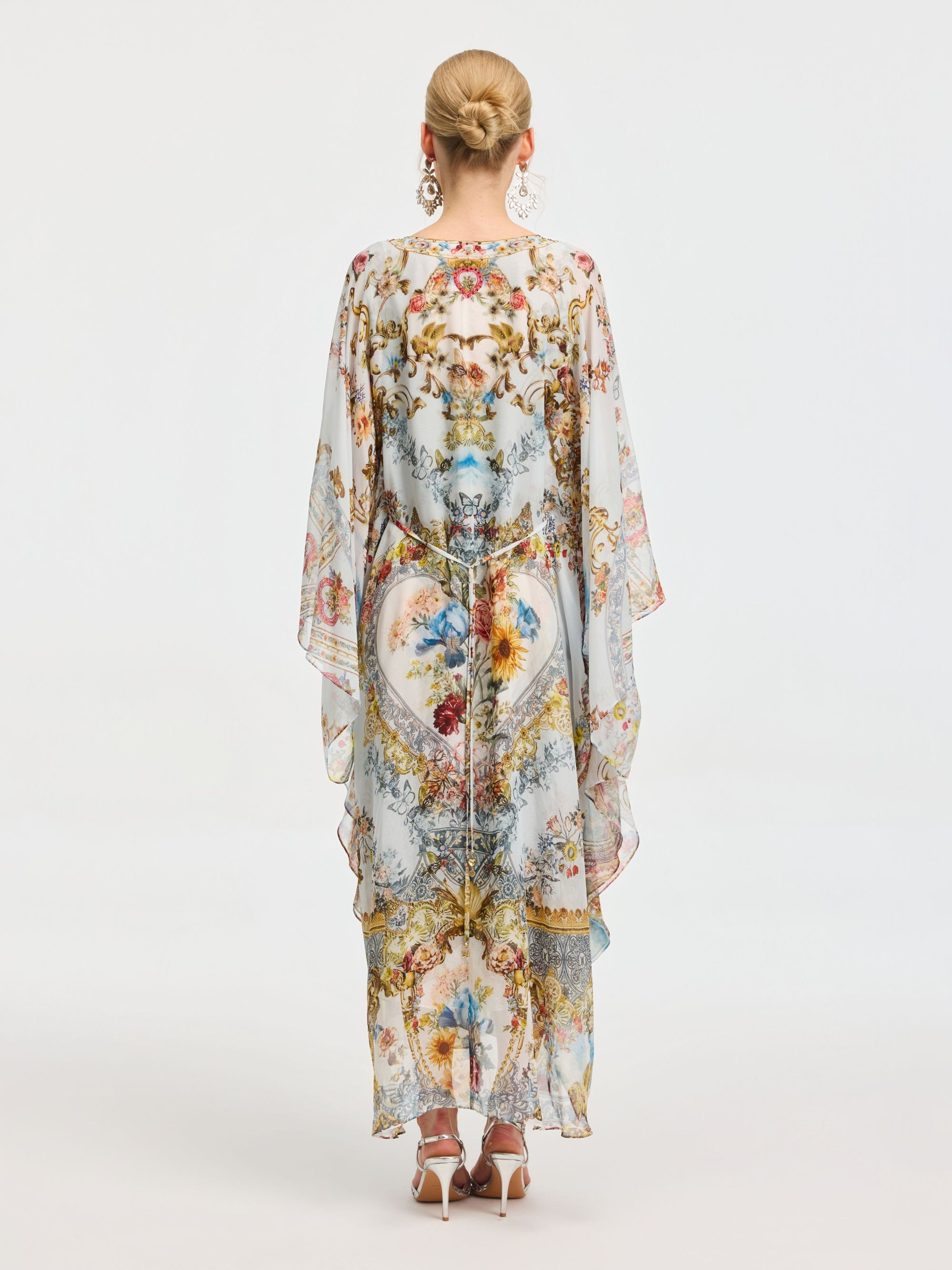 A woman with blonde hair in a bun is seen from the back wearing the Social Queen Baroque Floral Print Silk Caftan Dress, featuring wide sleeves and a long, sheer silhouette, set against a plain background.