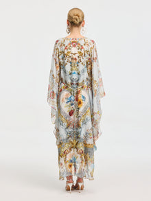 A woman with blonde hair in a bun is seen from the back wearing the Social Queen Baroque Floral Print Silk Caftan Dress, featuring wide sleeves and a long, sheer silhouette, set against a plain background.