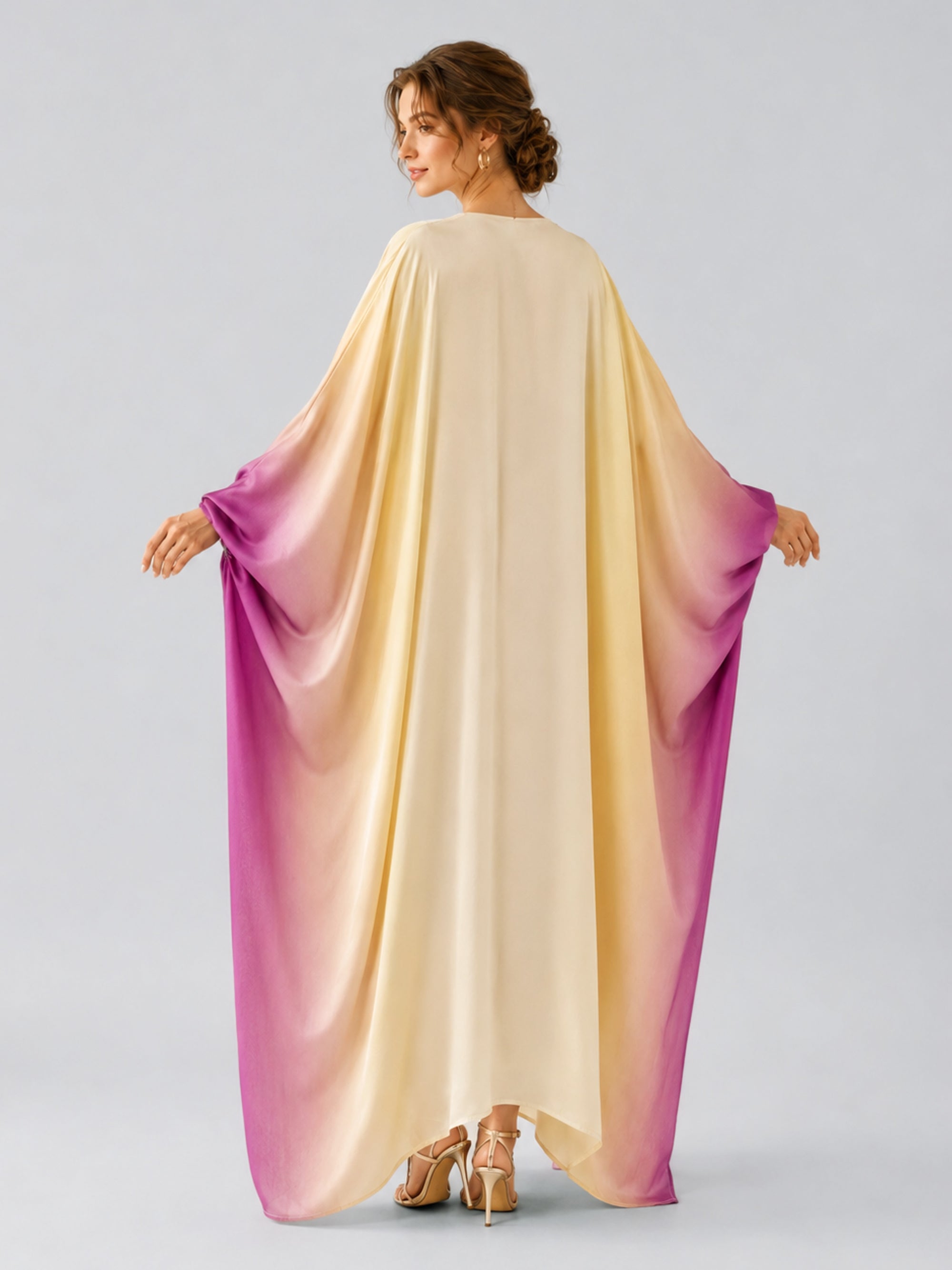 A woman stands with her back to the camera wearing the Social Queen Ombré Beaded Embroidered Cape Dress, which features a satin finish that fades from cream at the top to purple along the sleeves and hem, holding out the fabric to display its elegant drape.