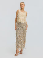 A woman stands facing forward against a plain light background, wearing the Social Queen 3D Leaf Beaded Mesh Top paired with strappy heeled sandals. The sleeveless beige top features delicate sequin fringe and intricate 3D leaf appliqués, creating subtle texture and visual interest in her ensemble.