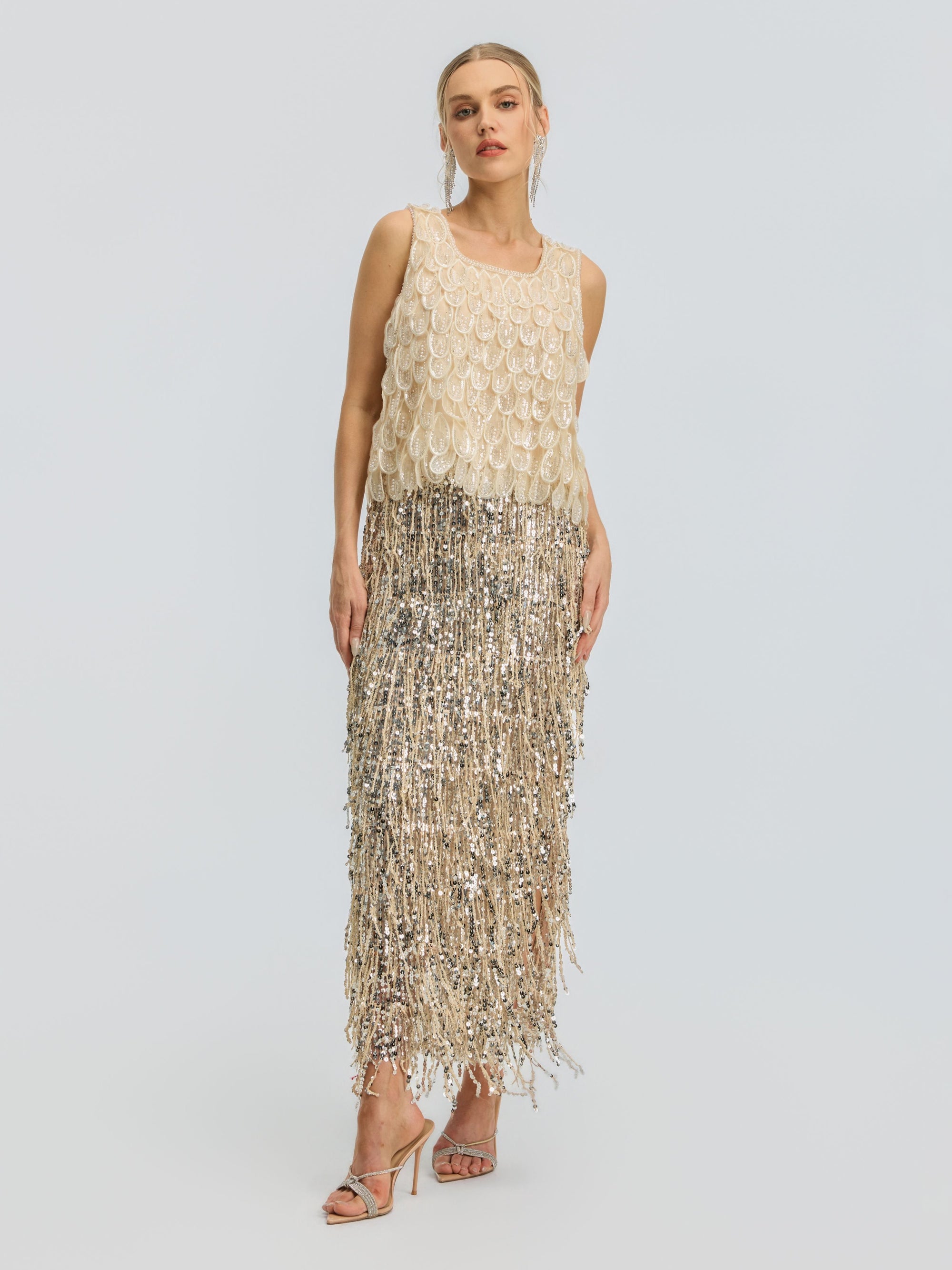 A woman stands facing forward against a plain light background, wearing the Social Queen 3D Leaf Beaded Mesh Top paired with strappy heeled sandals. The sleeveless beige top features delicate sequin fringe and intricate 3D leaf appliqués, creating subtle texture and visual interest in her ensemble.