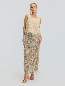 A woman stands facing forward against a plain light background, wearing the Social Queen 3D Leaf Beaded Mesh Top paired with strappy heeled sandals. The sleeveless beige top features delicate sequin fringe and intricate 3D leaf appliqués, creating subtle texture and visual interest in her ensemble.