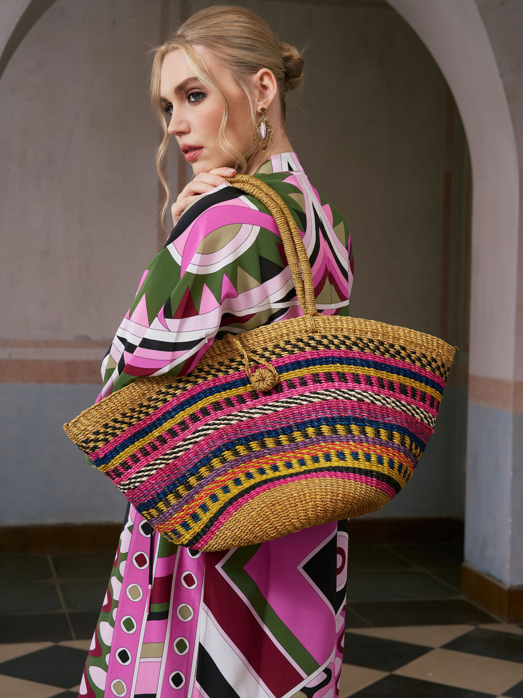 A woman stands indoors against an arched background, holding the Social Queen Colorful Striped Woven Tote Bag—a handwoven resort accessory crafted from natural ramie fiber and adorned with pink, yellow, and blue stripes.