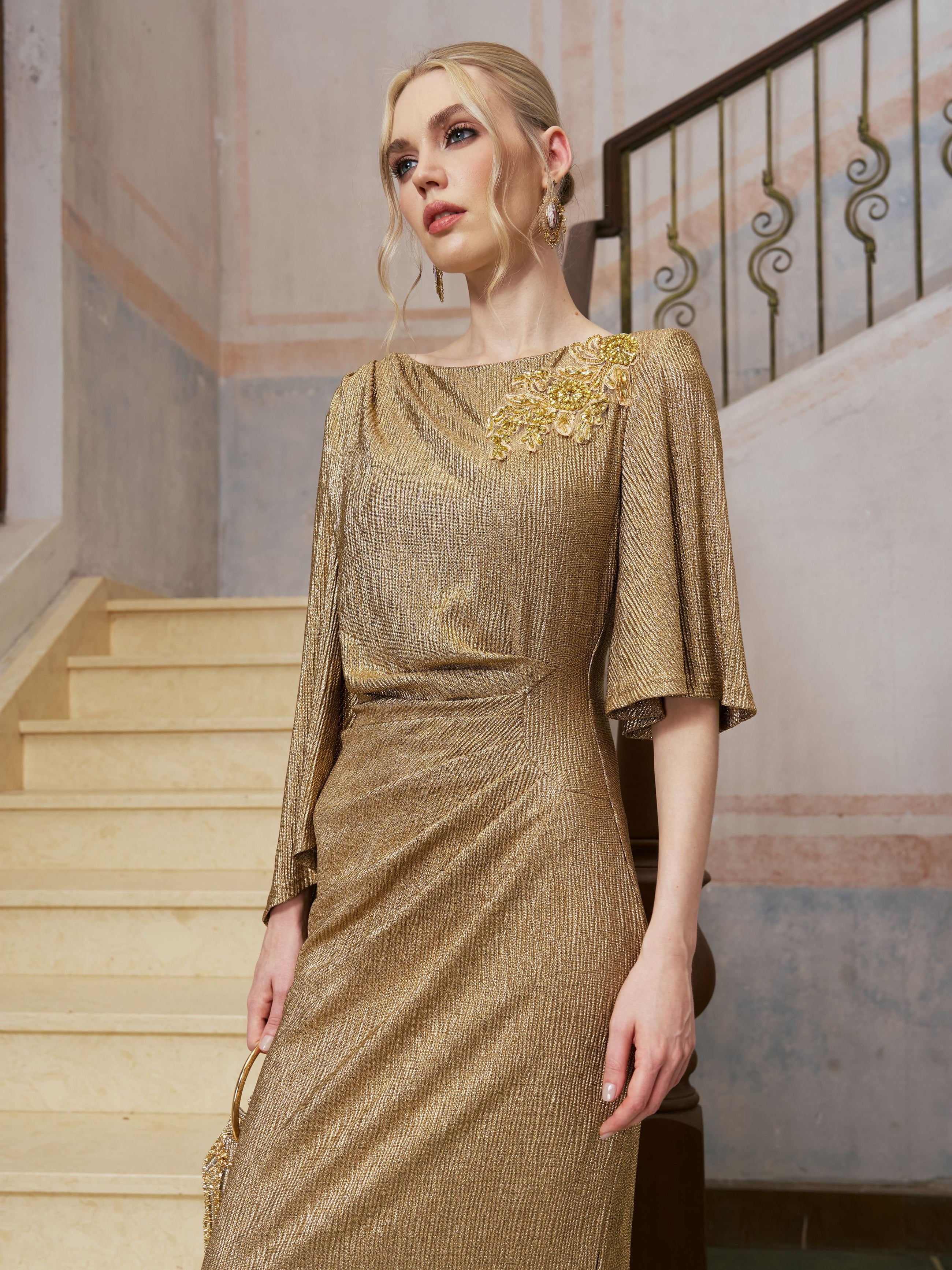 A woman wearing the Social Queen Floral Beaded Knit Dress, featuring gold metallic floral beadwork and intricate detailing, stands beside a staircase looking away from the camera.