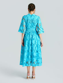 A woman faces away from the camera, wearing Social Queen's Rose Cutwork Lace Dress—a knee-length, bright blue A-line with rose-pattern lace and elbow sleeves—paired with white heeled sandals against a plain light background.