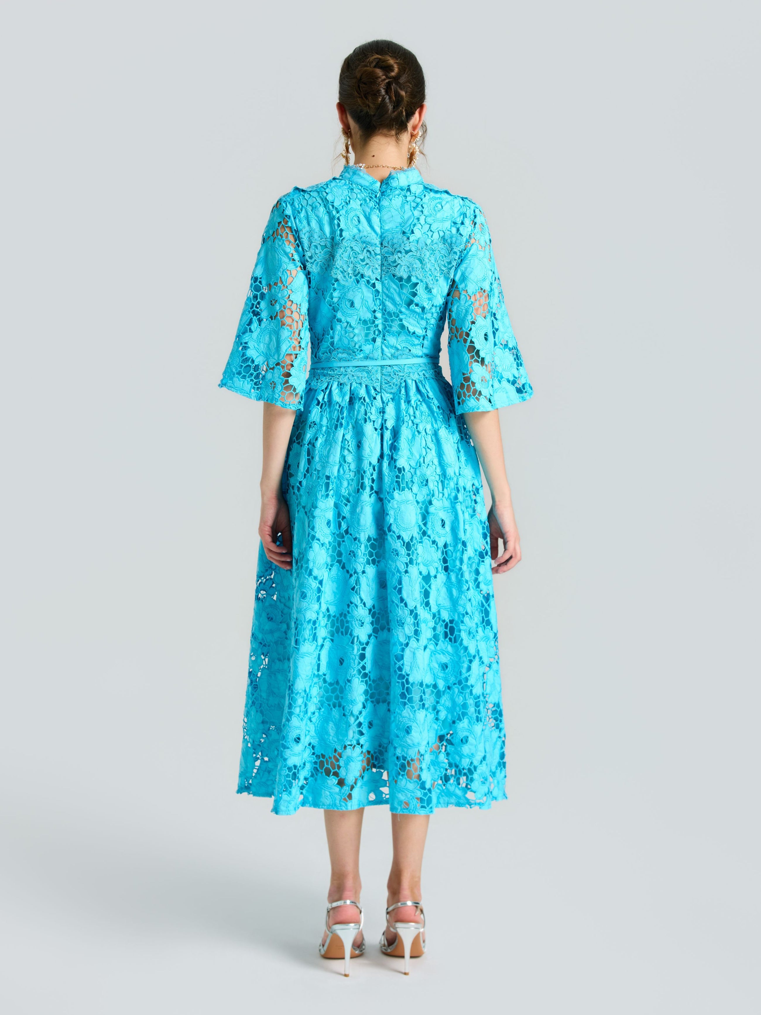 A woman faces away from the camera, wearing Social Queen's Rose Cutwork Lace Dress—a knee-length, bright blue A-line with rose-pattern lace and elbow sleeves—paired with white heeled sandals against a plain light background.