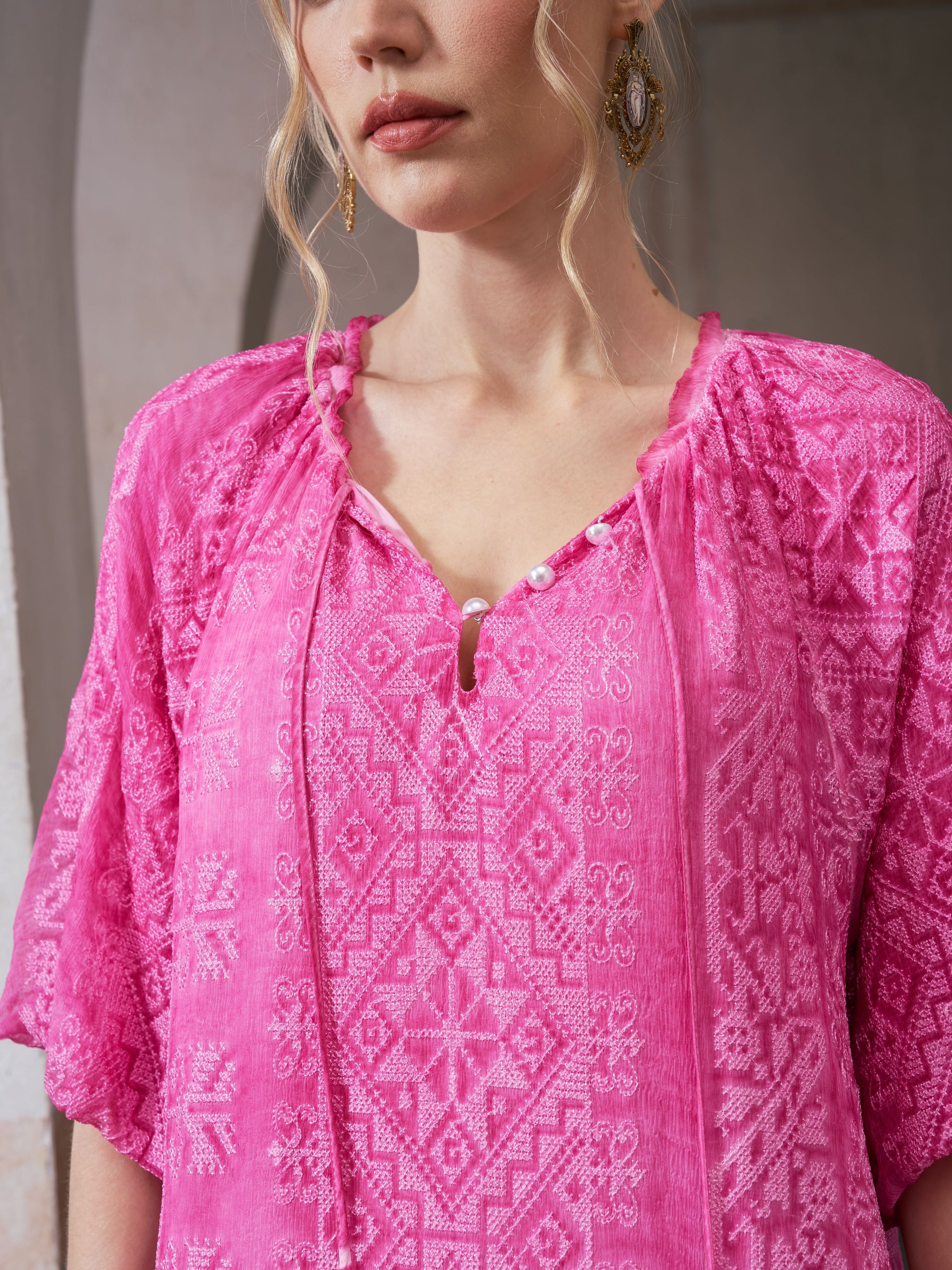 A woman with wavy blond hair and gold earrings wears the Social Queen Geometric Jacquard Mulberry Silk Dress in bright pink, standing indoors.
