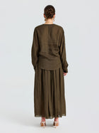 A person stands facing away, wearing a loose-fitting, long-sleeved olive blouse and the Social Queen Pleated Ramie Maxi Skirt (belt not included) with beige heeled sandals, set against a plain white background.