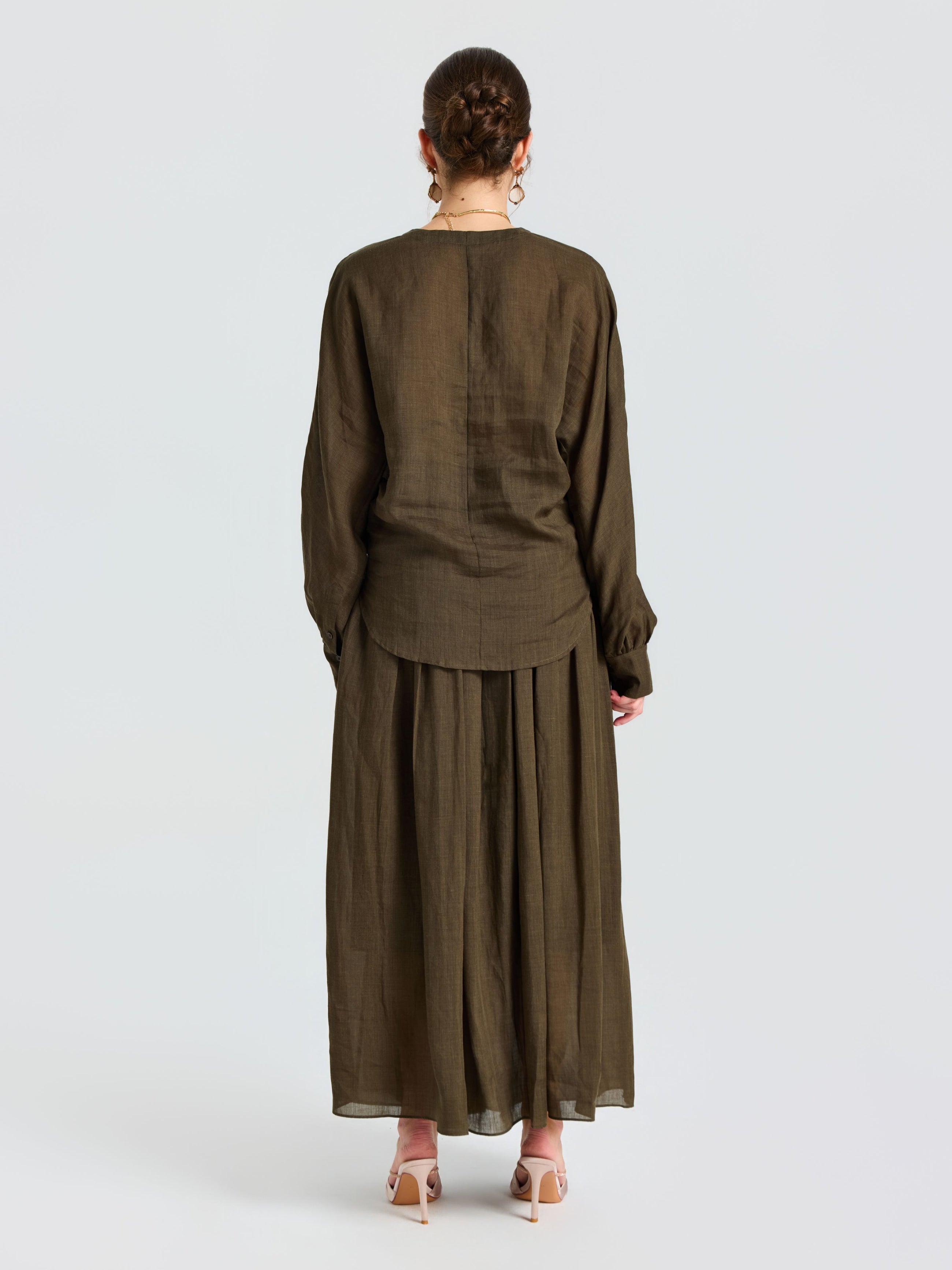 A person stands facing away, wearing a loose-fitting, long-sleeved olive blouse and the Social Queen Pleated Ramie Maxi Skirt (belt not included) with beige heeled sandals, set against a plain white background.