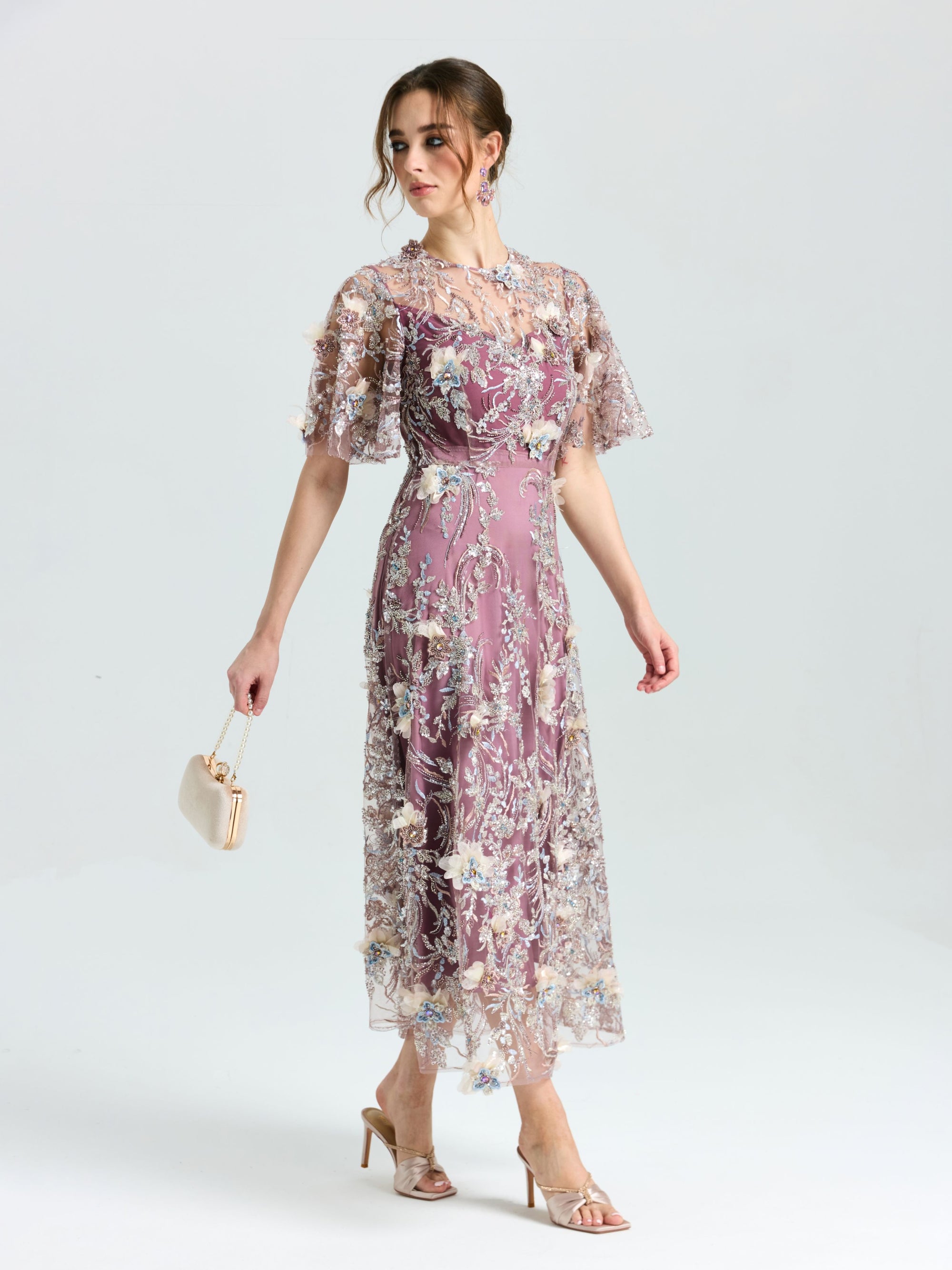 A woman in the Social Queen Floral Beaded A-Line Dress, featuring floral embroidery, sheer sleeves and hem, holds a small purse and wears high-heeled sandals against a plain background.