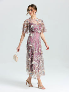 A woman in the Social Queen Floral Beaded A-Line Dress, featuring floral embroidery, sheer sleeves and hem, holds a small purse and wears high-heeled sandals against a plain background.