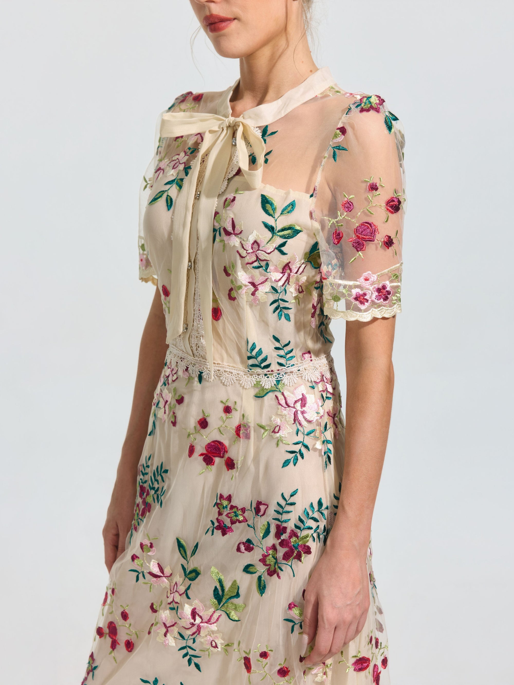 A woman models the Social Queen Floral Embroidered Bow-Tie Dress, a sheer mesh A-line dress with floral embroidery and a bow collar, against a plain background.