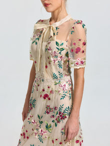 A woman models the Social Queen Floral Embroidered Bow-Tie Dress, a sheer mesh A-line dress with floral embroidery and a bow collar, against a plain background.