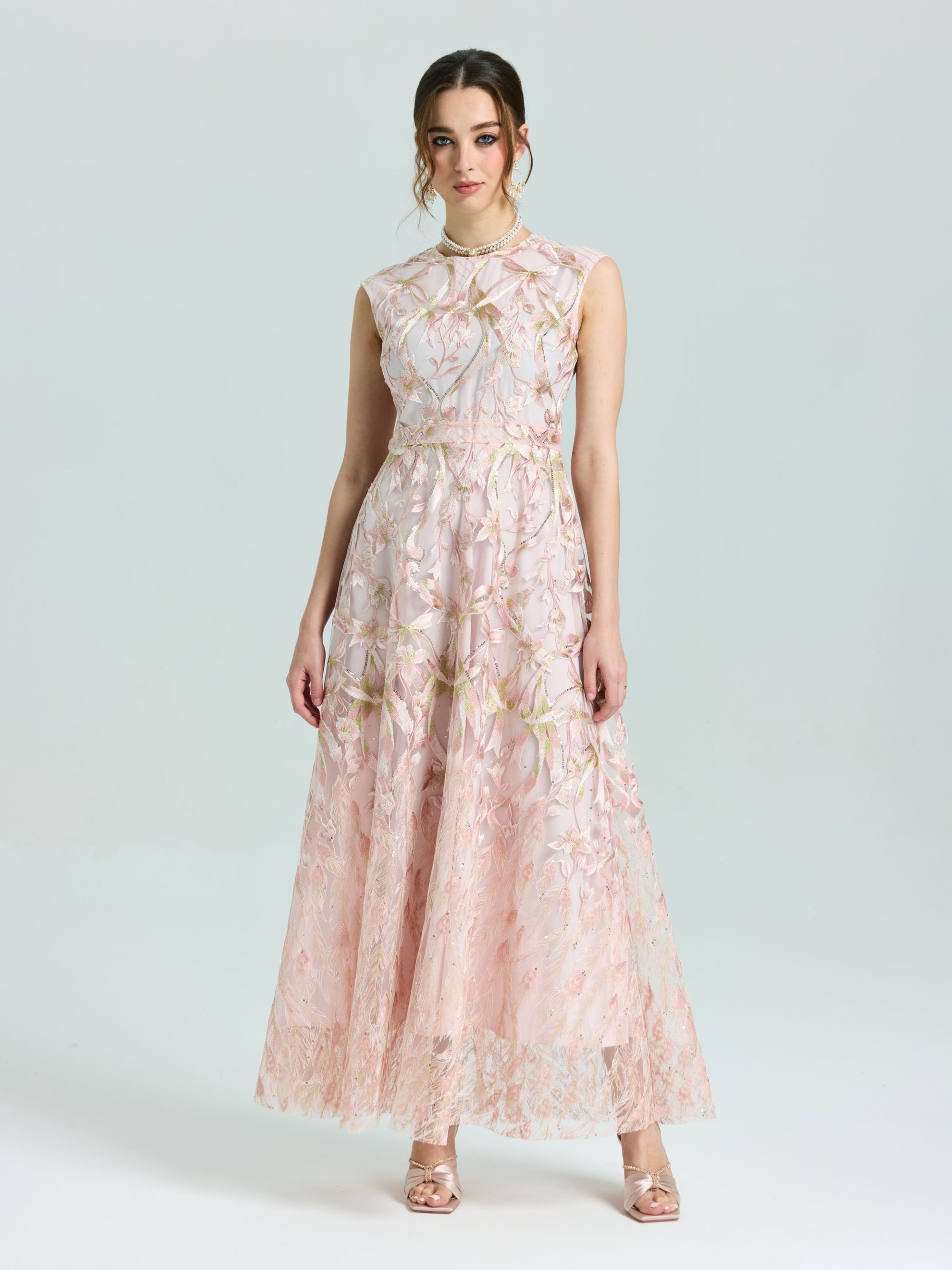 A woman stands against a plain background wearing the Social Queen Gold Floral Embroidered Dress, a sleeveless, floor-length A-line gown, paired with open-toe heels.