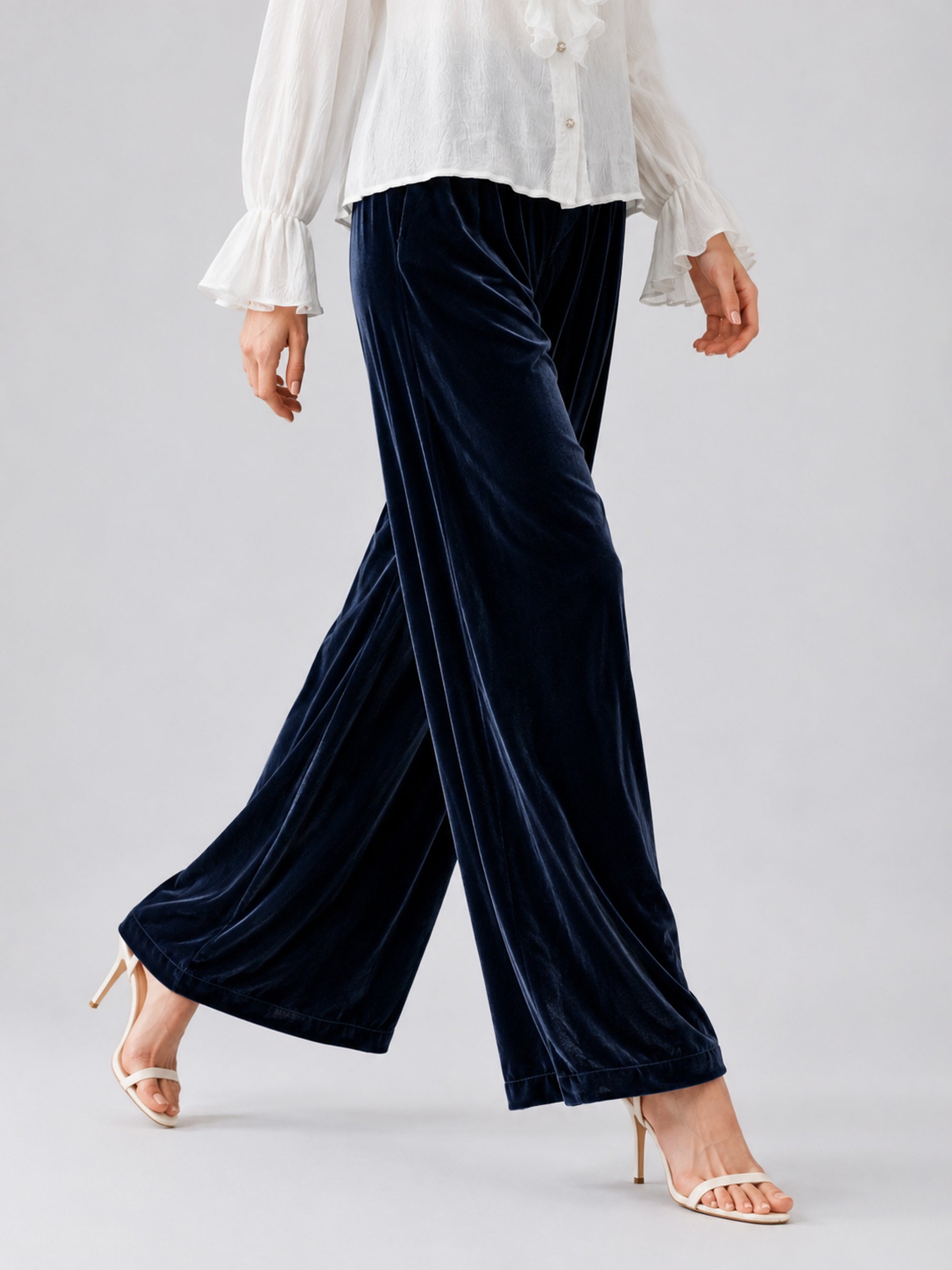 A person wearing a white long-sleeve blouse, Social Queen Silk Velvet Wide-Leg Pants in navy, and white heeled sandals, walking against a neutral background.