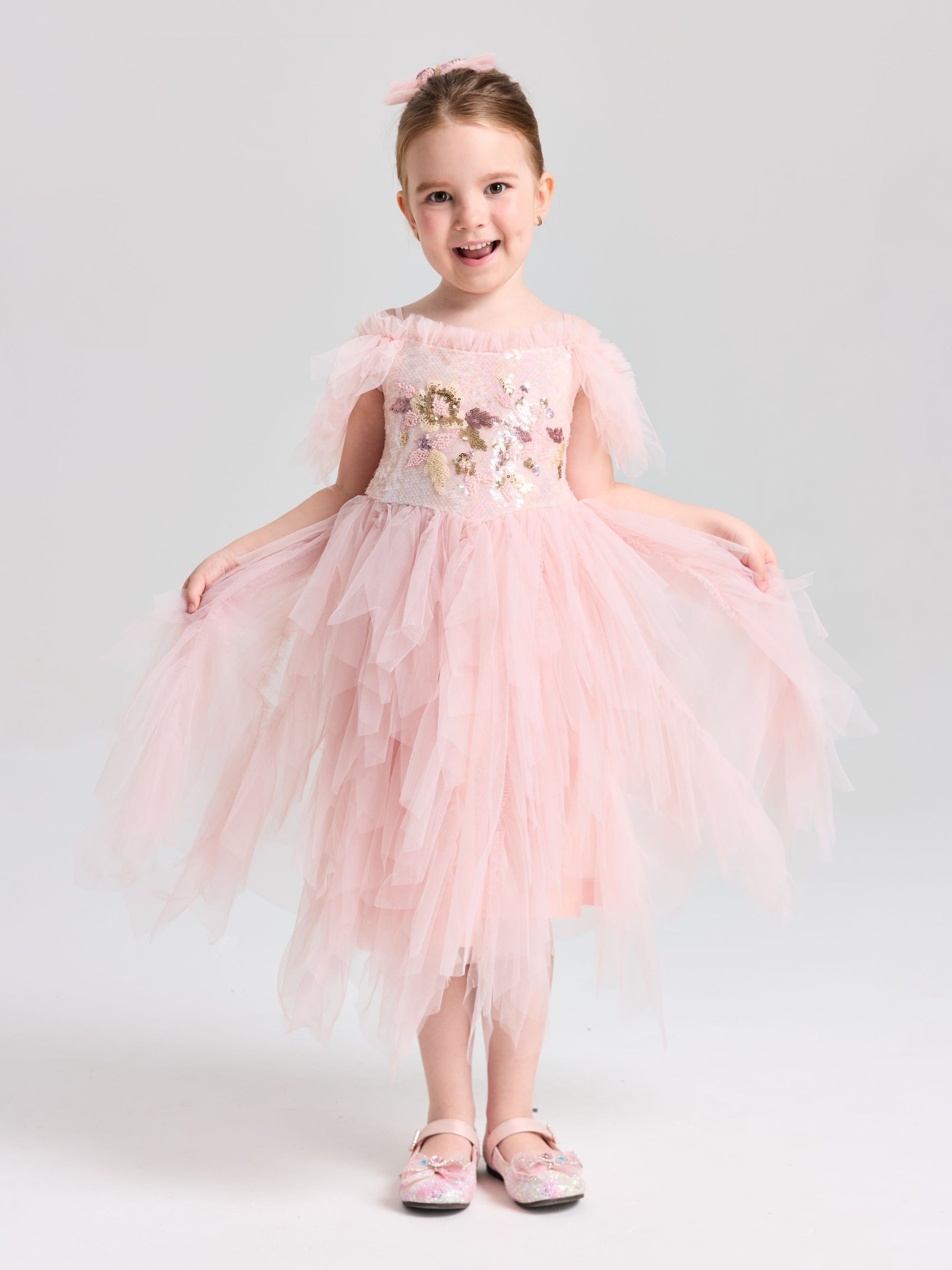 A young girl smiles in a Social Queen Girls Floral Sequin Tulle Dress, light pink and off-the-shoulder, paired with matching pink shoes.