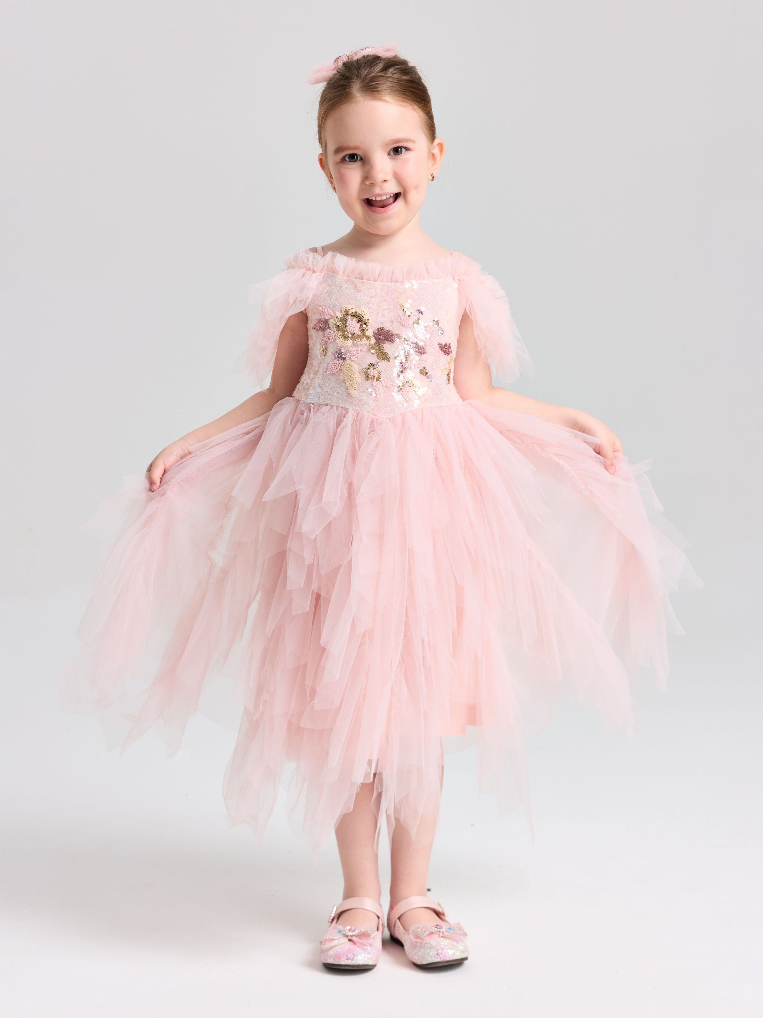 A young girl smiles in a Social Queen Girls Floral Sequin Tulle Dress, light pink and off-the-shoulder, paired with matching pink shoes.
