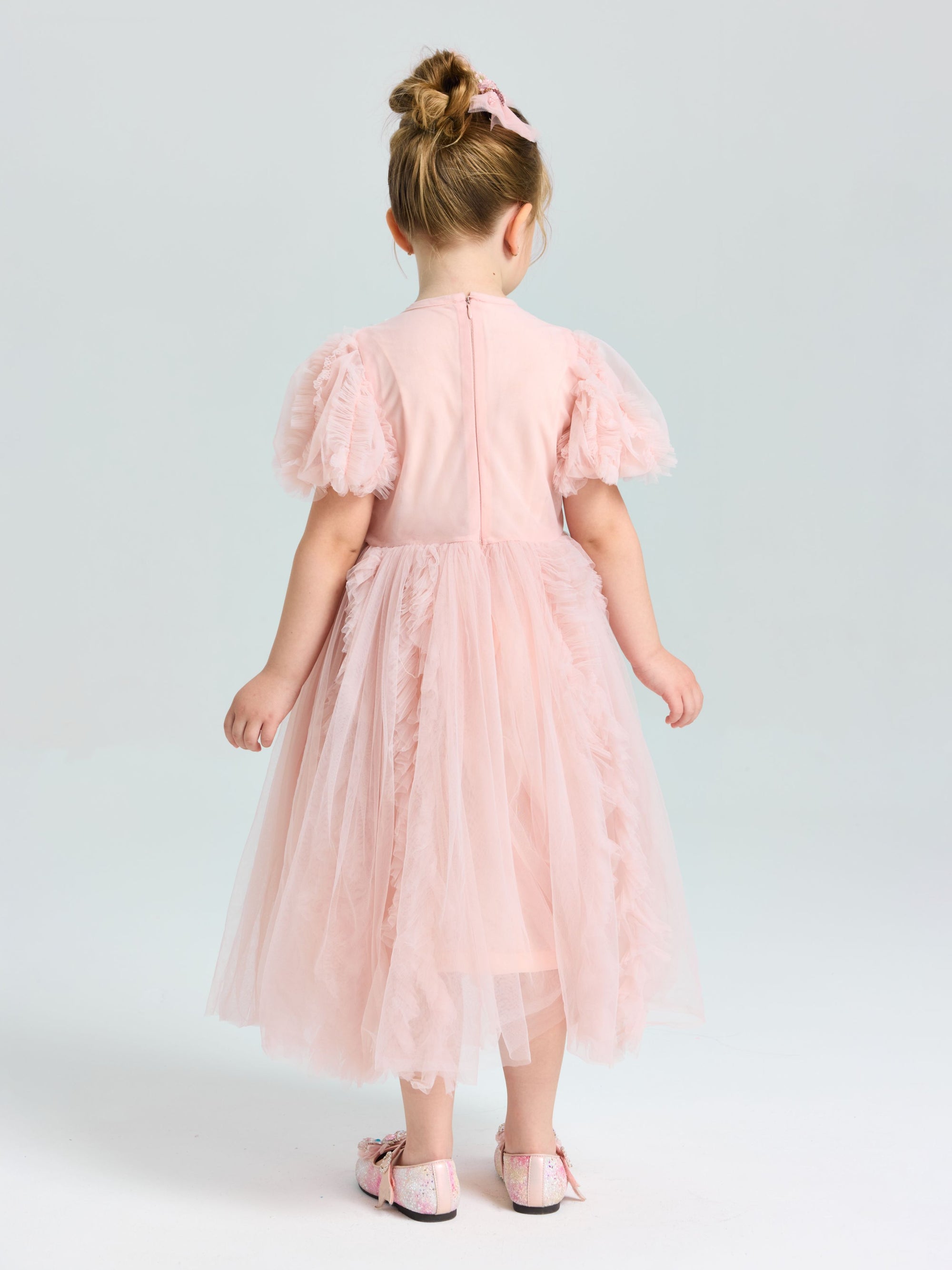 A young girl wears the Social Queen Girls Sequin Embellished Tulle Dress, featuring puff sleeves and a light pink, knee-length tulle skirt, as she stands facing away on a plain background for a dreamy look.