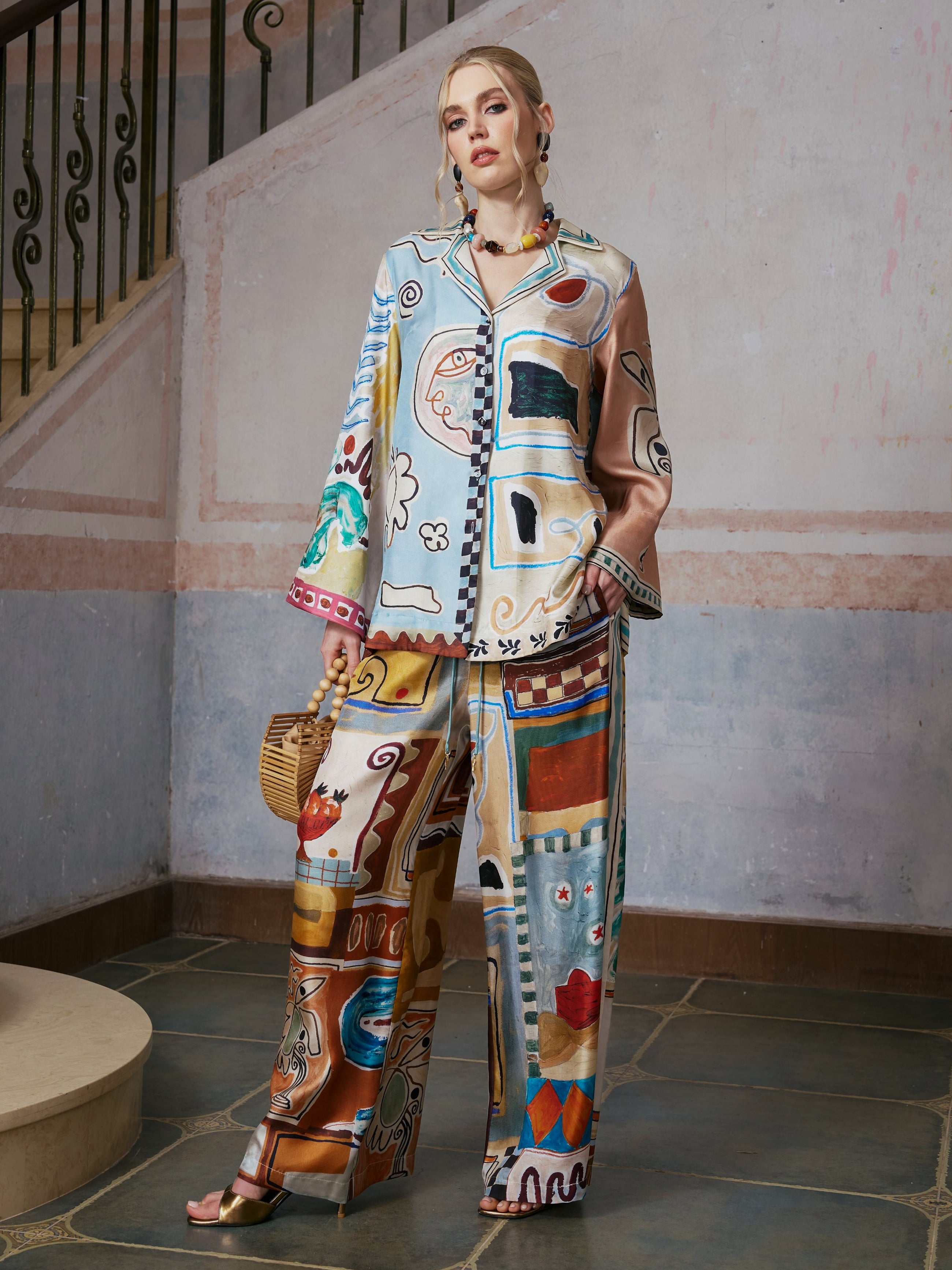 A woman stands indoors near a staircase, wearing the Social Queen Abstract Print Mulberry Silk Shirt as part of a matching set with loose resort wear. She accessorizes with gold sandals and holds a woven handbag.